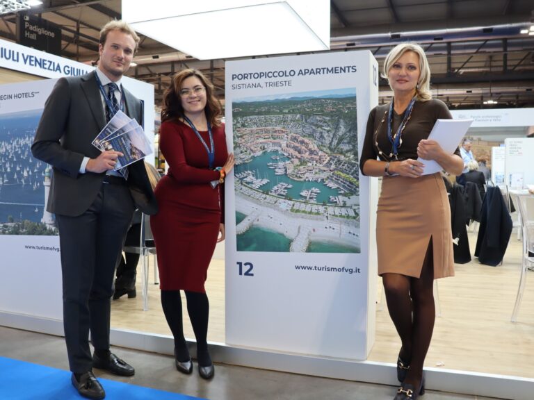 Tre persone in piedi accanto ad una colonnina con sopra scritto "Portopiccolo Apartments" ocn un'immagine aerea di Portopiccolo